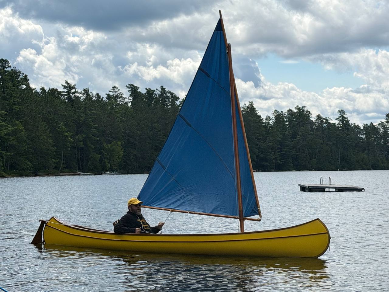 A yellow wood-and-canvas sailing canoe with a blue leg-o'-mutton sail on Big Wolf Lake, August 2026